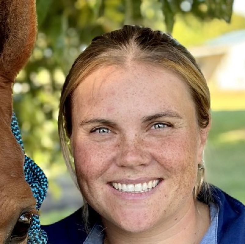 Ashleigh Fisher smiling next to a horse with a blue halter, outdoors in a sunny setting.