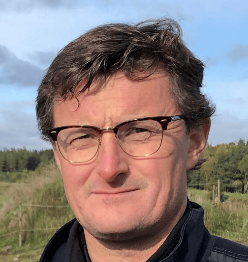 Dr. Lars Kristensen standing outdoors with a field and trees in the background under a partly cloudy sky.