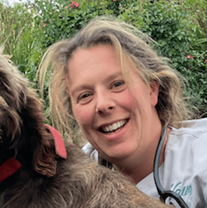 Dr. Libby Cooper smiling outdoors in a white veterinary uniform beside a brown dog with greenery in the background.