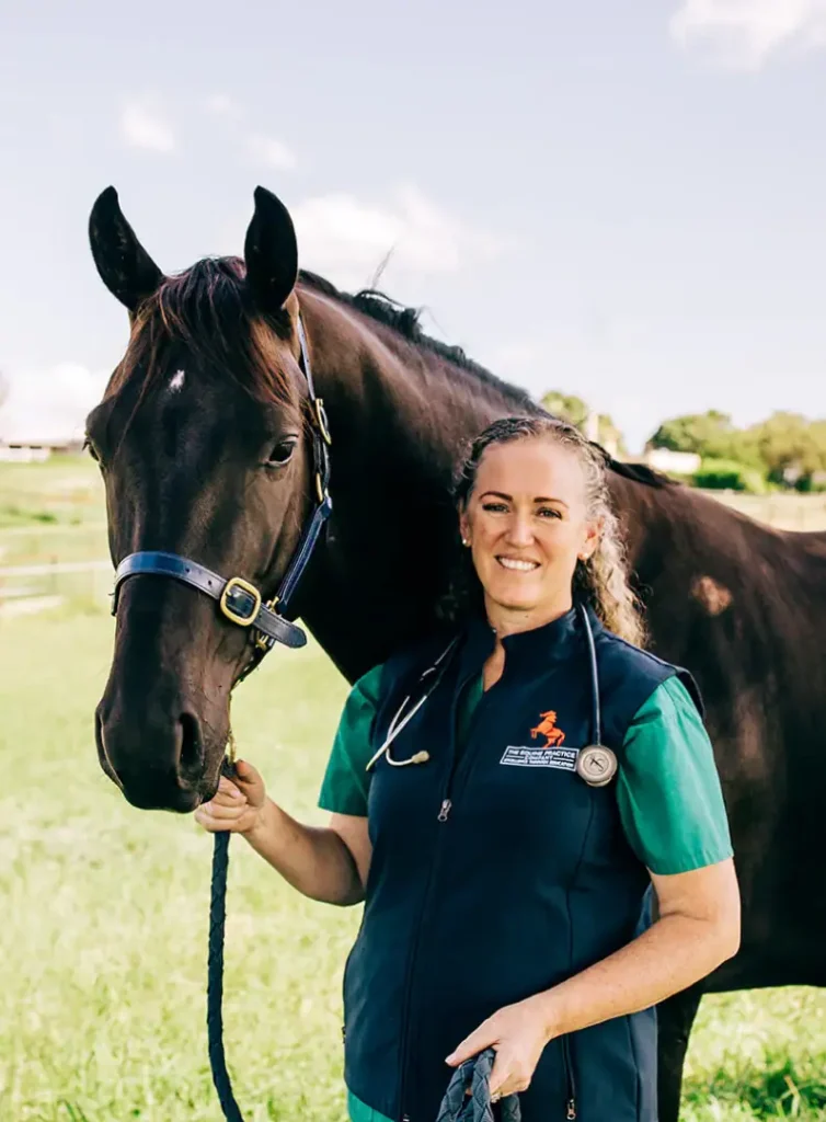 Dr. Olivia James, an equine veterinarian, standing beside a dark brown horse in a grassy field while wearing a stethoscope and clinic vest.